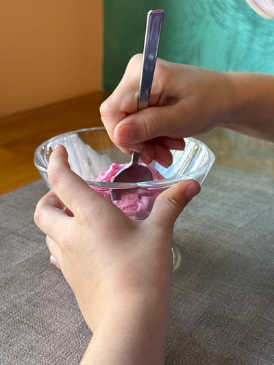 The image shows a close-up of a person's hands scooping pink ice cream from a clear glass bowl with a silver spoon. The person's hands and arms are visible, with one hand holding the spoon and the other holding the bowl. The background consists of a textured gray surface, possibly a couch or fabric. To the left of the hands, a bright green cloth is visible, and below that, a warm brown surface. The lighting suggests it is daytime, casting soft shadows. The scene appears to be indoors, in a casual setting, perhaps a kitchen or living room. There is no visible text in the image. The focus is on the action of scooping the ice cream, creating a sense of simple enjoyment or preparation for a treat.