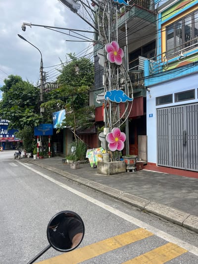The image shows a street scene in Quản Bạ Commune, Vietnam, on an overcast day. The foreground features the left side mirror of a motorcycle, reflecting the rider's dark shirt and a necklace. The street is paved with asphalt and has yellow and white painted lines, indicating a crosswalk. A concrete sidewalk runs along the right side of the street, with tactile paving for the visually impaired.
On the sidewalk, there is a pole with various decorations, including two pink floral shapes and blue cloud-like elements. A security camera is mounted on a pole overhead. To the right of the pole, there is a light blue building with a grey metal roll-up door and the number "52" affixed to the wall. Next to it, a darker building with red and green accents has windows and a balcony with a Mercedes-Benz logo.
Further down the street, on the left side, a motorcycle is parked near a blue building with large signage, including "HPT PH" and "AU". Trees and greenery are visible between the buildings, and other shops and structures line the street. A traffic sign with directional arrows is also present on the sidewalk. Several potted plants and some discarded items, including buckets and a wooden crate, are scattered along the sidewalk. The overall environment suggests a commercial or residential street in an urban setting.