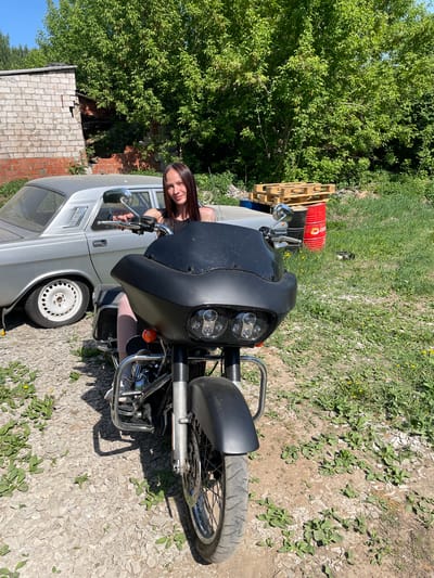 A young woman with long dark hair is seated inside a silver car with the driver's side window rolled down. A black motorcycle is parked directly in front of the car, obscuring part of the vehicle. The scene is set outdoors, with a ground cover of gravel and sparse vegetation. Behind the car, to the left, there is a small brick building. Further back, a lush green tree is visible. To the upper left of the car, wooden pallets and a red metal barrel are situated. The location is identified as Oktyabr'skii, Russia.