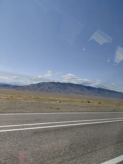 The image depicts a roadside view of a mountain range under a clear blue sky with scattered clouds. In the foreground, a paved road with two white lines running parallel is visible, suggesting movement or travel. The landscape transitioning from the road to the mountains is arid, with sparse vegetation, rocks, and gravel covering the ground. The mountains in the background are extensive and appear rugged, with some areas showing darker shadows and lighter, barren slopes. Reflections of what appears to be a vehicle's interior, including seats and possibly a window frame, are visible in the upper portion of the image, indicating the photo was taken from inside a vehicle. There are no people or animals clearly visible in the scene. The overall environment suggests a remote, possibly desert or steppe region. The location is identified as being in the city of Ынталы, Kazakhstan, which aligns with the arid, mountainous landscape.