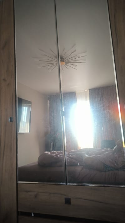 This is a vertical, medium-shot, eye-level photograph of a bedroom reflected in a large mirrored wardrobe. The wardrobe has a wooden frame and two mirrored doors. The reflection shows a bed with rumpled pink bedding, a window with brown curtains, and a ceiling light fixture that resembles a starburst. There is a television mounted on the wall to the left. Sunlight streams in through the window, creating a bright glare that obscures some details. The room appears to be lived in, with clothes draped over furniture. The overall impression is of a casual, domestic setting. The time of day appears to be morning or afternoon, given the strong sunlight. There is no visible text in the image. The location context is Kovrov, Russia.