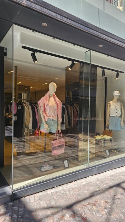 The image shows a retail store window display at street level in Charleroi, Belgium. The window is large and made of glass, allowing a view of the merchandise inside. The exterior features a black frame around the window and a dark, textured overhang above.
Inside the window, two mannequins are displayed. The mannequin on the left is wearing a pink t-shirt, a pink ruffled jacket, and light blue denim shorts. It is also holding a pink tote bag. A single white and grey sneaker is placed on a black mat below this mannequin. The mannequin on the right is wearing a white sleeveless top with buttons and a light blue skirt. It is holding a brown purse. To the right of this mannequin, a pair of yellow sandals and a small brown bag are displayed on a glass shelf.
In the background, racks of clothing are visible, suggesting the store sells apparel. The interior lighting consists of multiple spotlights mounted on a black track. Reflections of buildings and the sky are visible on the glass of the store window. The ground outside the store is paved with reddish-brown bricks, and sunlight casts shadows across the pavement.