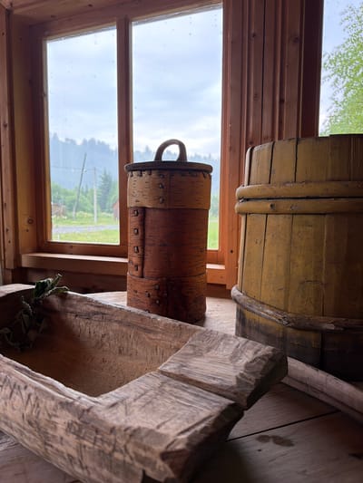 A still life composition is presented, featuring rustic wooden objects in an indoor setting with a view of the outdoors through a window. In the foreground, a weathered, trough-like wooden object is angled towards the viewer, its interior visible and containing a few dried green leaves. To the right of this trough, and slightly behind it, sits a cylindrical container made of birch bark with a dark lid featuring a wooden handle. Behind the birch bark container is a tall, cylindrical wooden barrel, painted yellow and showing signs of wear and tear with visible vertical planks and horizontal bands. The objects are arranged on a wooden surface, possibly a table or floor.
The background consists of a wooden-framed window looking out onto a natural landscape. The window pane is slightly clouded or wet, obscuring some details of the view, which includes a treeline of coniferous trees, some distant buildings, and a sky that appears overcast. The interior framing of the window and the wall to its right are made of unfinished wood planks. The lighting is natural, coming from the window, and casts soft shadows. There are no people or animals visible in the scene. The overall impression is one of rustic simplicity and a connection to traditional craftsmanship. The image is believed to be taken in Ust'-Pyzas, Russia, given the context.