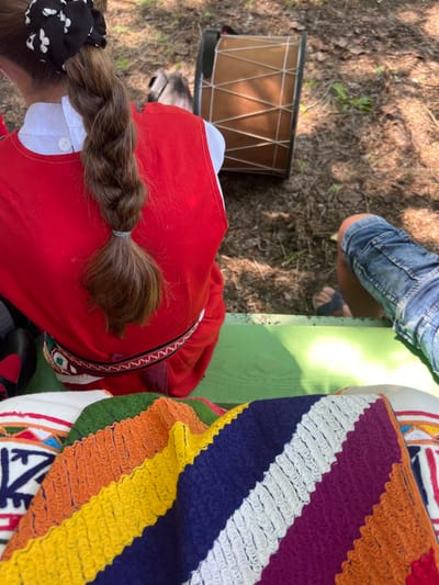 The image depicts an outdoor scene in Bolyarovo, Bulgaria, viewed from a slightly elevated perspective. In the central foreground, a person is observed from the upper back, wearing a bright red sleeveless garment over a white collared shirt with a visible white button near the left shoulder. Their medium brown hair is styled in a long, thick braid, secured at the top with a black scrunchie patterned with white flowers and at the end of the braid with a light grey elastic band. Around the waist, a decorative woven belt with geometric patterns in red, black, and white is visible.
To the left of the central figure, a black shoe with a red accent is partially visible. Further left, a dark garment suggests the presence of another individual. To the right of the central figure, the lower right leg of a third person is visible, clad in blue denim shorts and a dark-colored sandal on their foot.
The individuals appear to be seated or positioned near a bright green painted wooden surface that occupies the lower portion of the frame. Draped over this green surface in the immediate foreground is a vibrant, multi-colored knitted or crocheted blanket with distinct diagonal stripes of green, orange, yellow, blue, white, and purple, exhibiting a textured, ridged pattern. To the left of this blanket, a white textile with embroidered or woven patterns in blue, red, and orange is partially visible.
In the background, behind the individuals, the ground consists of sun-dappled dirt and scattered dry leaves, indicating a natural, outdoor environment. A rectangular, light brown object, appearing to be a drum with white crisscrossing strings on its side, rests horizontally on the ground.
