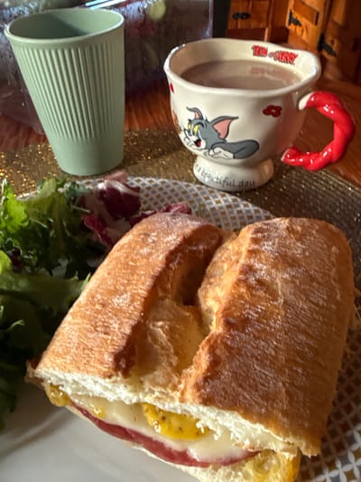 A partially eaten sandwich rests on a plate amidst a salad of mixed greens. The sandwich reveals layers of cheese, salami, and mustard between slices of crusty bread. Nearby is a Tom & Jerry themed mug filled with a light-colored liquid, and a green, ribbed tumbler. The scene is shot at an angle, set upon a shiny, reflective surface.