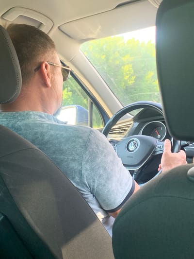 The image depicts the interior of a vehicle, likely a Volkswagen, based on the emblem on the steering wheel. A male driver, wearing sunglasses and a light blue textured shirt, is seen from behind, holding the steering wheel. His right hand is visible on the wheel, with his thumb extended upwards in a possible gesture. The dashboard shows a speedometer with a needle indicating a low speed. Outside the car, through the windows and windshield, a sunlit environment with lush green trees and foliage is visible, suggesting a rural or natural setting. The overall scene is captured from the perspective of a passenger seated in the back of the vehicle. The image does not contain any information that would allow for the identification of the location as Rakhmatova Polyana, Russia.