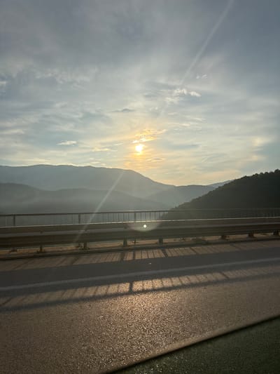 The image depicts a sunset over a mountainous landscape, viewed from a bridge. The sun is partially visible, casting rays of light through a hazy sky. The mountains are layered, with the farthest ones appearing in a soft, bluish silhouette, while closer ones are darker and more defined. A guardrail runs horizontally across the foreground, separating the road from the precipice. The road surface, made of asphalt, reflects the warm light of the setting sun, creating a shimmering effect. Shadows of the guardrail are cast onto the road. The scene suggests a tranquil and scenic viewpoint. The location is identified as the area around Vrachesh, Bulgaria.