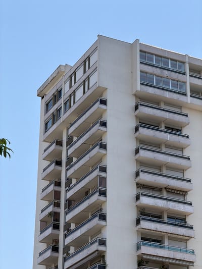 A low-angle medium shot captures a portion of a tall, modern residential building in Courbevoie, France, set against a clear, pale blue sky. The building features a repetitive architectural style with numerous balconies jutting out from its facade. These balconies are uniformly designed with white railings and dark trim, some equipped with retractable awnings. The windows on the building appear to be covered by roller shutters, giving the facade a consistent, somewhat uniform appearance. The concrete and stucco surfaces of the building show some signs of wear and tear, with visible discoloration and minor damage, particularly along the top edge. A few green leaves from an unseen tree or plant are visible in the upper left corner, adding a small touch of nature to the otherwise urban scene. The building's structure is angular and geometric, reflecting a mid-to-late 20th-century architectural aesthetic.