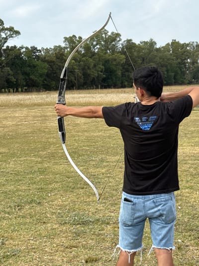 A person in a black shirt and denim shorts holds a recurve bow. The bow's string is taut. The person is outside, standing on a grassy field, with trees in the background. The sky appears cloudy. The bow is silver and black. The shorts have frayed edges. The shirt has a design on the back.
