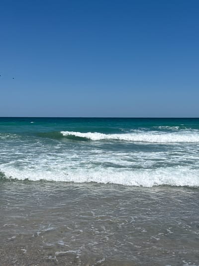 This image depicts the coastline of Shabla, Bulgaria, on a clear day. The scene is dominated by a vast expanse of calm, bright blue sky that takes up the top half of the frame. Below the sky, the deep turquoise waters of the sea stretch horizontally across the image. Several rows of white-capped waves are visible, with the closest ones gently lapping at the shore. The water appears clear and the seabed is visible through the shallow water near the foreground, composed of small stones and sand. Two small black birds are present in the sky, near the top left of the frame. The overall impression is one of tranquility and natural beauty.