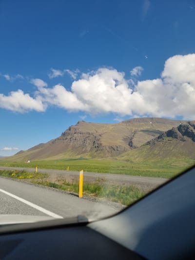 The image depicts a scenic landscape with a prominent mountain range in the background, under a bright blue sky with scattered white clouds. In the foreground, a road runs parallel to a grassy field dotted with yellow wildflowers. Yellow posts mark the side of the road. The perspective suggests the photo was taken from inside a vehicle, with part of the car's interior visible in the lower portion of the frame. A distant white building with a red roof is visible in the field to the left of the mountains. The scene appears to be a rural or semi-rural area outside of a city. Given the description of Iceland and the landscape, the location is likely on the outskirts of Reykjavik, perhaps along a scenic route.