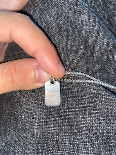 The scene, located in Lawrence Township, United States, depicts a close-up view of a human hand holding a small, rectangular silver-colored pendant. The pendant, attached to a thin, silver-colored chain with a heart-shaped bail, displays etched Hebrew text: "יברכך ה' וישמרך." The hand holds the pendant against a dark grey, textured fabric surface, likely clothing. No other individuals or environmental features are discernible.