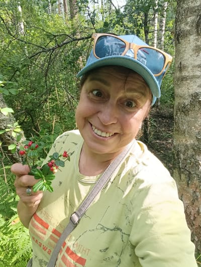 A middle-aged woman with brown hair and fair skin is taking a selfie outdoors. She is wearing a light yellow t-shirt with a graphic print, a blue baseball cap, and large sunglasses with wooden frames perched on the cap. She is smiling broadly and holding a small sprig of red berries with green leaves in her left hand. The background is a forest with green foliage, trees, and sunlight filtering through the leaves. The woman is positioned in the foreground, with a tree trunk on the right side of the frame. The scene appears to be a candid outdoor shot, possibly taken during a walk or hike in Vasiutino, Russia.