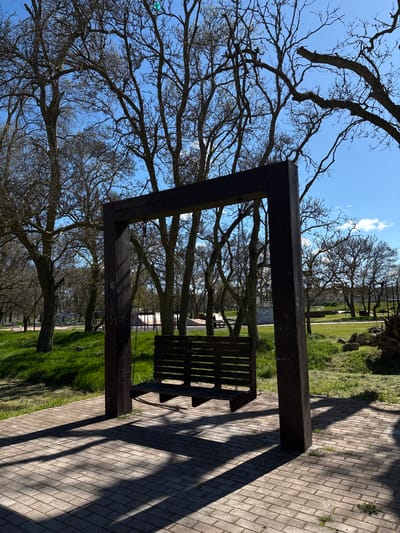 The image shows a wooden bench with an attached dark brown rectangular frame, situated on a brick walkway in a park. Sunlight casts strong shadows from the bench and frame onto the walkway. Behind the bench and frame, there's a view through the frame to trees with bare branches against a blue sky. Green grass is visible on either side of the walkway, and more trees and structures are visible in the background. The location is Chornomorskoe, Ukraine.