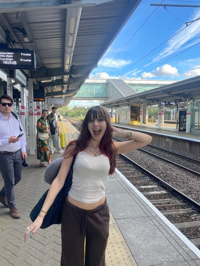 This is a medium shot photograph taken from a train platform in Luton, United Kingdom. The perspective is slightly below eye level and angled upwards towards the subject in the foreground. The subject, a young woman with long, dark hair with red highlights, is wearing a white tank top and brown sweatpants. She is facing the camera with her mouth open in an excited expression and her right arm is raised, resting on her head. She is wearing several rings on her left hand and bracelets on her right wrist. A dark bag is slung over her left shoulder.

In the middle ground of the photograph, there are several other individuals on the platform. To the left, a man in a white shirt and sunglasses is walking away from the camera, holding a mobile phone and a black object. Behind him, a woman in a floral dress is looking to her left. Further down the platform, another person is visible. A yellow suitcase is positioned on the platform.

The environment is a train station platform with a covered waiting area supported by pillars. Train tracks run alongside the platform. Overhead, electrical wires are visible against a bright blue sky with scattered white clouds. In the background, a modern pedestrian bridge crosses over the tracks. A digital display board above the platform shows information about train departures, including "Sandwich [Kent]", "Three Bridges", and times such as "19:21:35". Another sign indicates "Luton Airport Parkway". The platform itself is paved with textured tiles.