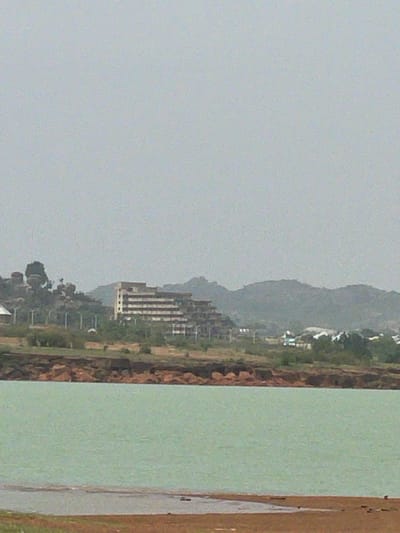 The image shows a landscape of Nabatum, Nigeria on an overcast day. A body of water is on the right side of the frame, bordered by a brown shoreline. The central part of the image contains a stretch of land with sparse vegetation, including trees and low-lying plants. A high-rise building stands out against the backdrop of distant hills, suggesting the presence of urban or developed areas within the natural scenery. The sky is uniformly gray, indicating a cloudy or foggy weather condition.