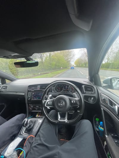 The image depicts the interior of a Volkswagen car, specifically the dashboard, steering wheel, and center console area, as viewed from the driver's perspective. The car is being driven on a road lined with trees. A road sign is visible in the distance. The driver is wearing dark clothing, and part of their leg is visible. A pack of cigarettes is visible next to the gearstick.
Based on the road signs, foliage, and car model, it can be inferred that the location is likely within the city of Bolton, United Kingdom.