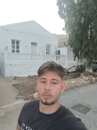 This is a medium shot, selfie-style photo of a young man with short, light brown hair and a light beard. He is wearing a dark gray t-shirt with white text on it. He is looking directly at the camera with a neutral expression.

The man is standing in front of a white, two-story house with multiple windows and doors. A white railing runs along the front of the house. To the right of the house, there is a large tree. In the background, a dark-colored car is parked. There is also a small garden tiller or tilling machine on the ground near the house. The scene appears to be outdoors during daylight, likely late afternoon or early evening given the soft lighting. The ground is paved, and there is a stone wall in the middle ground. The overall setting suggests a residential area, possibly in a warm climate. The text on the man's shirt is partially visible and appears to be an English phrase, with "BACK TO" clearly readable. The municipality of Chalki, Greece, is the stated location context, suggesting a Greek island setting.