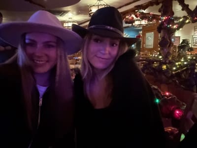 Two women in cowboy hats, one in dark blue and the other in light gray, smile at the camera. They appear to be indoors, possibly in a restaurant or bar decorated with holiday lights and rustic wooden accents. The image is taken at an unusual angle, as if the viewer is on the ceiling looking down. A glimpse of an American flag and framed artwork is visible in the background.