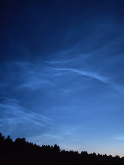 This is a photograph taken at twilight in Volokov Most, Russia. The image features a dark silhouette of a dense forest lining the bottom portion of the frame. Above the treeline, the sky is a deep, gradient blue, transitioning from a darker hue at the top to a lighter shade towards the horizon. Wispy, luminous clouds, possibly noctilucent clouds, are scattered across the sky, appearing bright white against the deep blue backdrop. The overall scene conveys a sense of tranquility and natural beauty during the evening hours.