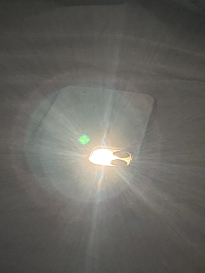 The image displays an overhead perspective of an interior space, presented as located in Angwan Sayawa, Nigeria. A prominent, intensely bright light source is positioned centrally, emitting strong white-yellow light rays that create a starburst lens flare effect across the frame. This main light fixture features a light-colored, possibly metallic, housing with an irregular shape and a distinct decorative cutout on its right side. To the immediate left of the primary light, a smaller, square-shaped, vibrant green light source is visible. The dominant background is a uniform, medium-grey, textured surface, consistent with a ceiling or upper wall, which exhibits subtle lighter variations. No individuals, additional distinct objects, or specific actions are discernible within the frame.