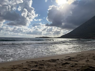 The image depicts a beach scene in Vlichada, Greece, with the ocean in the foreground and a dark, mountainous landscape in the background. The sky is dramatic, filled with a mix of light and dark clouds, with the sun shining through, creating a bright, shimmering effect on the water. Waves are breaking on the shore, leaving white foam on the dark sand. Several small figures, likely people, are visible in the water, some appearing to be surfing or engaging in water sports. The overall impression is one of a wild and beautiful natural setting.