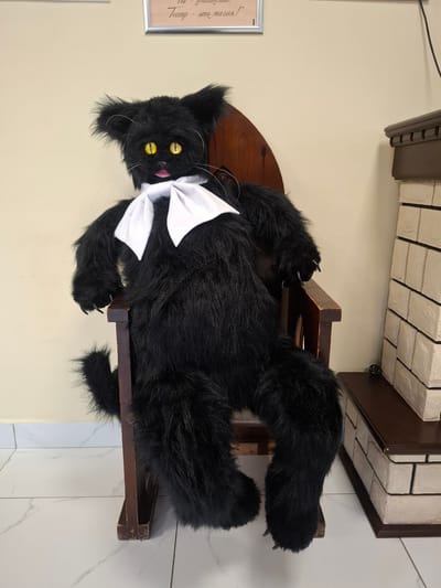 A plush black cat doll with yellow eyes and a white bow sits on a dark wooden chair. The doll has long, shaggy fur and large paws with visible claws. It is positioned in front of a pale yellow wall. To the right of the doll is a decorative fireplace with a brown mantelpiece and a facade made of light-colored bricks. A framed picture hangs on the wall above the doll. The floor is tiled with a white marble pattern. The scene is indoors, likely in a public space given the framed text on the wall, possibly a theater or cultural center in Baykalsk, Russia.