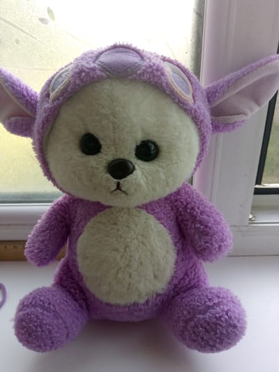 A stuffed teddy bear, predominantly purple with a cream-colored belly and face, is positioned in the foreground. The bear is wearing a purple hood with large, floppy ears and appears to be sitting on a white surface, possibly a windowsill. Behind the bear, a window with a translucent pane is visible, allowing diffuse light to illuminate the scene. The window frame is white. To the left of the bear, a portion of a wooden ruler with markings is present. A thin purple string or yarn lies on the white surface near the bear's left foot. The environment suggests an indoor setting with natural light coming from the window. The location is identified as Prigorodnoye, Kyrgyzstan.