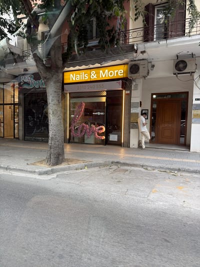 The image depicts a street scene in Thessaloniki, Greece. In the foreground, a mature tree trunk is prominently featured on a sidewalk, with its branches extending into the upper portion of the frame. Behind the tree, a commercial establishment with a bright yellow awning displays the signage "Nails & More" with a phone number below it. The storefront has large windows, one of which showcases a floral decoration spelling out "Love." To the left of this establishment, another storefront with a dark, rolled-down shutter is visible. On the right side of the image, a man wearing a light-colored shirt and pants is standing by a wooden door, looking at his phone. The building facade above the storefronts displays air conditioning units. The street and sidewalk show signs of wear, with some debris and markings on the pavement. The overall impression is of a typical urban commercial street.