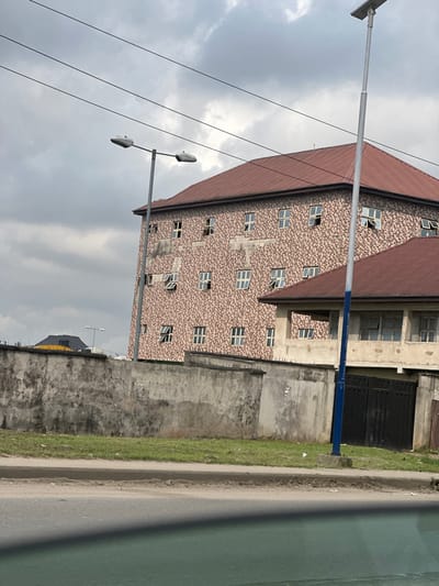 The image depicts an urban street scene in Rumuogonoma, Nigeria, under an overcast sky. In the foreground, a grey asphalt road runs horizontally, bordered by a strip of green grass and a weathered concrete perimeter wall. This wall, a light grey, shows signs of cracking and discoloration, and features concrete coping and some pointed security elements along its top edge.
Behind the wall, two streetlights are visible. To the left, a standard streetlight with a grey pole supports two horizontal lamp fixtures. Above it, three parallel power lines traverse the frame. To the right, a taller, slender streetlight with a blue base section features a solar panel-equipped lamp at its apex.
The background is dominated by a multi-story building, likely an apartment complex or commercial structure, featuring a prominent terracotta-colored hip roof. Its facade is covered with small, textured tiles in a pattern of beige, brown, and off-white tones. Numerous rectangular windows with white frames are distributed across its visible four stories, with many windows partially or fully open. Areas of exposed, lighter-colored concrete are visible where tiling is absent or incomplete on the building's facade. To the right and slightly in front of this main building, a smaller, two-story structure with a similar terracotta hip roof is visible. This secondary building, appearing lighter in color, includes several windows and a dark, possibly metal, gate at ground level. A distant building with a dark roof is faintly visible in the far left background, beyond the main wall. No individuals are present, and no active interactions or movements are captured.