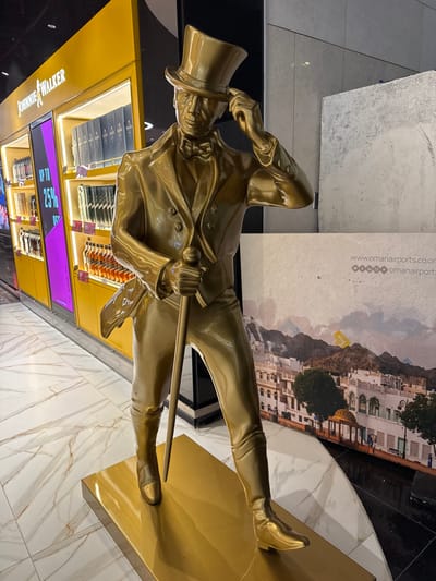A golden statue of the Johnnie Walker striding man stands in front of a duty-free store. He wears a top hat and holds a cane, embodying a confident stride. The store features backlit displays with bottles, and a purple electronic sign shows "UP TO 25%". In the background is a picture of a town, possibly a tourist destination, and the floor has a marble pattern.
