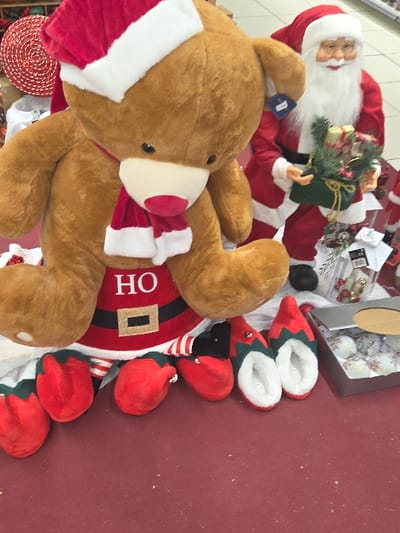 This image captures a festive retail display in Blainville-sur-Orne, France. The central subject is a large, brown plush teddy bear wearing a red and white Santa hat, a red scarf, and a red apron designed like a Santa suit, featuring "HO" in white text and a gold-buckled black belt. To the right and slightly behind the teddy bear stands a Santa Claus mannequin, dressed in a red suit with white trim, a white beard, and glasses, holding a green fabric sack filled with wrapped gifts and green foliage. In the foreground, positioned on a red floor, are several pairs of red and green elf-style slippers with white fleece interiors. To the right of the slippers, a gray rectangular box holds multiple white spherical Christmas ornaments, some with snowflake designs. The background reveals a partial view of a red spiral decoration on the left and a brightly lit retail environment with white tiled flooring visible behind the Santa mannequin.