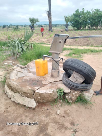 In Angwan Hausawa, Nigeria, a scene unfolds around a manual water pump. The pump, made of weathered metal, stands on a cracked concrete base, its lever arm extended. Adjacent to it, a bright yellow jerrycan rests, likely filled with water. Two black tires are stacked on the ground near the pump, one positioned on top of the other, with a piece of concrete lodged between them. A thin black rope is tied around the pump's support and the upper tire. The environment is dusty and arid, with sparse green grass growing around the base of the pump and tires. In the background, palm trees and other foliage dot a field, suggesting a rural or semi-rural setting. Two figures, possibly children, are visible in the distance, one in red and another in darker clothing, walking across the field. The sky is overcast with clouds. The bottom of the image displays a watermark reading "Samsung Galaxy A03".