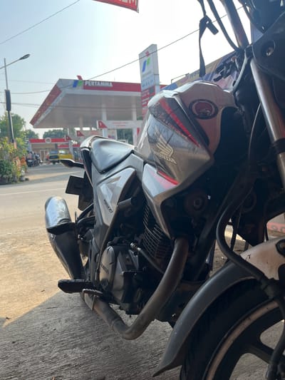 The image shows a motorcycle parked in front of a Pertamina gas station in Boja, Indonesia. The motorcycle is a Honda, with visible branding on the fuel tank and engine. The Pertamina station has its distinctive red and white branding, with pumps visible in the background. The scene appears to be on a street, with a road and sidewalk present. The lighting suggests it is daytime, with the sun casting shadows. There are other vehicles and people in the distance, indicating a typical street scene in a populated area. The focus is on the motorcycle in the foreground, with the gas station and its surroundings serving as the backdrop.