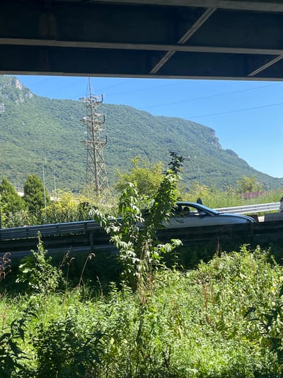 A silver convertible car is seen driving on a road with a metal guardrail, framed by lush green vegetation in the foreground and a tree-covered mountain in the background. A power line tower is visible on the mountainside. The scene appears to be near Lecco, Italy.