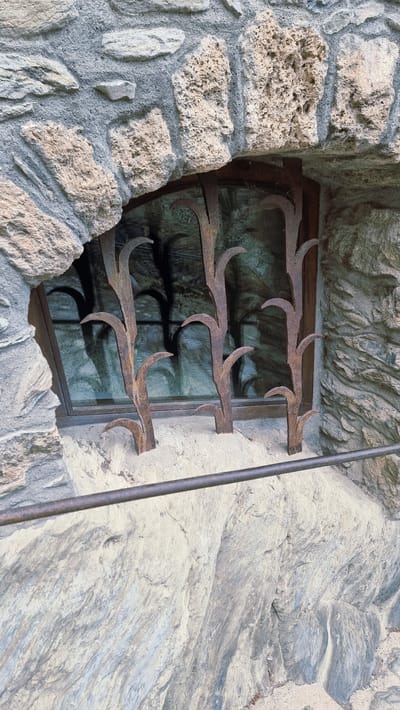 The image shows a close-up view of a rustic stone window. The window opening is irregular and framed by rough-hewn, light-colored stones with visible texture and some pitting. The stones are mortared together with a greyish material. Inside the opening, a dark-colored windowpane reflects the surrounding environment, showing a shadowy, arched pattern. In front of the window, a decorative wrought-iron grill is visible. The grill is made of dark, rusted metal and features stylized vertical elements that resemble stalks of corn or wheat, with curved leaf-like protrusions. The base of the grill appears to be set into a sandy or fine-grained aggregate material. A horizontal metal bar, also appearing to be rusted, runs across the lower portion of the image, in front of the stone structure and below the window. The overall impression is one of age and a natural, rugged aesthetic. This window is likely located in Camprodon, Spain, a town known for its medieval architecture and stone constructions.