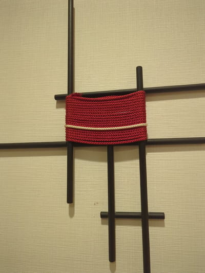 The image depicts a close-up view of a wall decoration. The background is a textured, light-colored surface, possibly wallpaper, with a subtle horizontal lining pattern. Superimposed on this surface are several dark, cylindrical rods arranged in a cross-like configuration. A prominent feature is a thick, tightly woven red cord wrapped around the intersection of these rods. A thin, lighter-colored cord, possibly white or cream, is also wrapped around the red cord, accentuating its texture. The lighting casts soft shadows, suggesting an indoor setting. The composition is minimalist and appears to be a decorative element within a space. This artwork is located in the city of Narita, Japan.