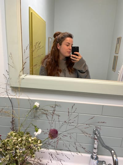 An indoor scene depicts a mirror selfie taken in what appears to be a washroom or bathroom in Kaiserslautern, Germany. The primary subject is a female individual reflected in a rectangular mirror with a light-colored frame. She has long, wavy reddish-brown hair, partially gathered in a top bun, and is wearing a distressed grey knit sweater. She holds a black smartphone with her right hand, capturing the image, and a silver ring is visible on her left ring finger. Her gaze is directed towards the phone screen.
In the reflected background, a light blue-grey wall is visible. To the left, a portion of a pale yellow door or panel is reflected. To the right, two small framed items are mounted vertically on the wall. Below the mirror's reflection, a section of light blue-grey subway tiles is present.
In the foreground, a floral arrangement partially obscures the bottom left of the mirror and the view of the sink area. This arrangement consists of dried-looking green and beige hydrangea-like clusters, two white carnation-like flowers, one spherical purple flower (likely an allium), and numerous stalks of dried grasses with small seed heads. To the right, a white porcelain sink basin is partially visible, along with a polished chrome faucet featuring a lever handle. The overall lighting is soft and even.