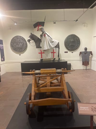 In a museum setting, a life-sized diorama depicts a knight on horseback, dressed in chainmail and a white surcoat with a red cross, facing to the left. The horse is also adorned with a surcoat bearing a red cross. Behind the horse, a black and white flag with a red cross is displayed. Flanking the horse on the wall are two large, circular metal plaques, each with intricate embossed designs. To the right of the horse, a mannequin is dressed in medieval chainmail armor. In the foreground, a wooden siege weapon, identified by a nearby sign as a "Mangonel Attack device," is positioned on a black platform. The museum floor is tiled with reddish-brown stones. The overall impression is a historical exhibit focused on medieval warfare. The image was taken in the city of Toledo, Spain, at the Museo de los Ejércitos.