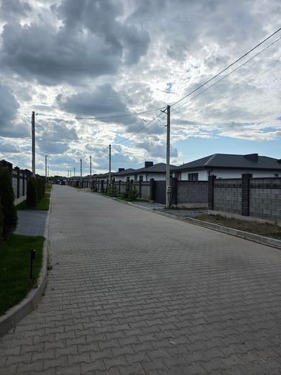 The image depicts a street in the city of Velykyi Zhytyn, Ukraine, lined with modern houses and fences. The street is paved with interlocking grey bricks, and a sidewalk runs along the left side, bordered by a curb and grass. On the left, there is a row of manicured evergreen trees. The houses, characterized by white walls and dark grey tiled roofs, are separated by fences constructed from grey concrete blocks and dark metal gates. Several utility poles with overhead wires are visible along the street, and the sky above is filled with dramatic, heavy grey clouds interspersed with patches of blue sky. A single black car is parked on the street in the distance. The scene appears to be a residential area, likely a new development.