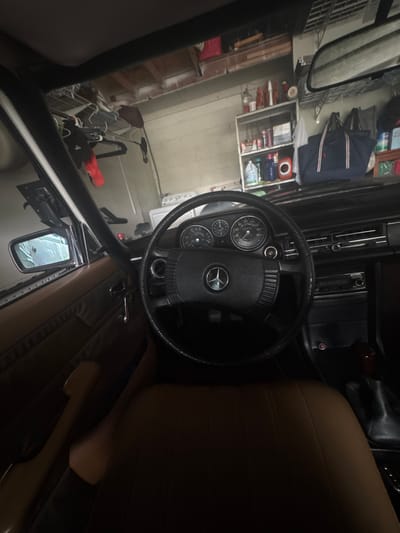The image provides a driver's perspective from inside a Mercedes-Benz vehicle, stationary within a residential garage in Miami Shores, United States of America. The vehicle's interior features a black four-spoke steering wheel with the Mercedes-Benz emblem, an analog instrument cluster behind it, a dark-colored dashboard, and brown/tan upholstered seats and door panels. A wooden-topped gear shifter is visible on the center console to the right. The garage environment beyond the windshield displays light-colored walls, consistent with concrete or painted block, and an exposed wooden beam ceiling structure. On the left side of the garage, a white large appliance, possibly a washing machine, is present. A metal shelving unit against the rear wall is stocked with various bottles, containers, and miscellaneous items. Above this shelving, multiple bags, including a prominent navy blue duffel bag with red and white stripes, are stored or hung. Additional clothing items, including a red garment and a dark jacket, hang from hooks or a rack on the left wall. No individuals are depicted, and no active interactions are occurring. The scene is illuminated by ambient, diffuse light.