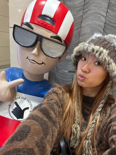 A young woman with light skin and long, light-brown hair poses beside a statue. She wears a furry hat and a brown-and-black striped sweater. The statue has a red-and-white striped helmet and sunglasses. It also has a logo with a hand pointing, labeled "Vintage The Hot Pot." They stand in front of a gray tiled wall, creating a selfie-style photograph.