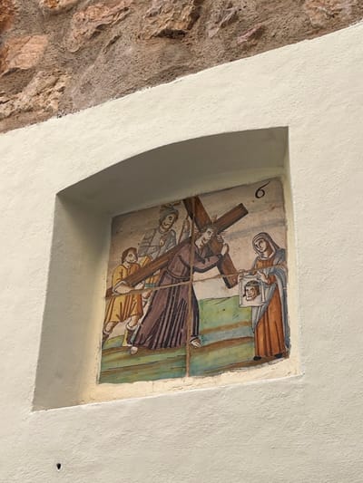 An arched niche in a textured, cream-colored plastered wall in Fornalutx, Spain, houses a tile mural depicting a religious scene. Above the plaster, a section of an exposed stone wall is visible. The mural, identified by a numeral "6" in its upper right corner, illustrates the Sixth Station of the Cross. In the center, a man, likely Jesus, wears a crown of thorns and struggles under the weight of a large wooden cross carried on his right shoulder. He wears a dark, long-sleeved robe. To his immediate left (viewer's right), a woman in a blue head covering and an orange dress holds up a white cloth bearing the imprinted face of a man. Behind the central figure, on the left side of the mural, a soldier in a helmet and armor pulls the cross via a rope. To the soldier's left, another man in a yellow tunic appears to assist by supporting the cross from below. The ground beneath the figures is depicted in horizontal bands of green and brown. The mural exhibits visible lines, suggesting it is composed of multiple painted tiles. A small dark hole is visible in the lower left section of the cream-colored plaster wall surrounding the niche.