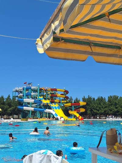 The image depicts a sunny day at a water park in Khudat, Azerbaijan. In the foreground, a portion of a striped yellow and grey awning is visible, casting a shadow over the scene. The background is dominated by a large, multi-colored water slide complex with blue, white, yellow, and red sections, towering above a clear blue swimming pool. Several children are visible in the pool, some swimming and others floating on inflatable rings. Red and yellow inflatable buoys are scattered across the water. A table with a straw bag, a water bottle, and some snacks is in the lower right corner. Trees line the perimeter of the water park, and a clear blue sky completes the scene.
