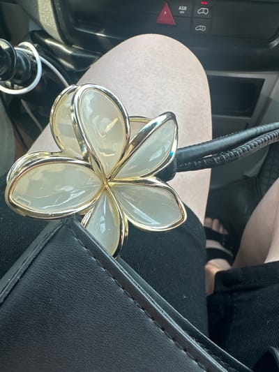 In Chomentowo, Poland, a close-up shot focuses on a person's lap in a vehicle. The person is wearing black shorts and has bare feet visible. Resting on their lap is a black handbag and a gold-colored flower-shaped accessory with translucent white petals. The background shows a car's dashboard with buttons and a warning light.