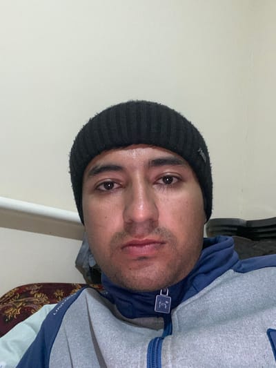 An adult male subject is depicted in a front-facing portrait, located indoors in Galaosiyo, Uzbekistan. The subject has a light to medium skin tone, dark eyes, and displays light facial stubble. He is wearing a black ribbed knit beanie and a two-tone zippered jacket; the main body is light grey, while the collar and shoulder sections are dark blue. A blue zipper is visible, with a square, white-on-dark logo zipper pull. The subject maintains a neutral facial expression, looking directly towards the camera. The background consists of a plain, light-colored wall. A white horizontal pipe or rail is partially visible on the left side of the frame, behind the subject. Below the pipe, a dark fabric with a light, intricate pattern is visible. A dark, undefined object is partially visible on the far right. Lighting appears to be artificial.