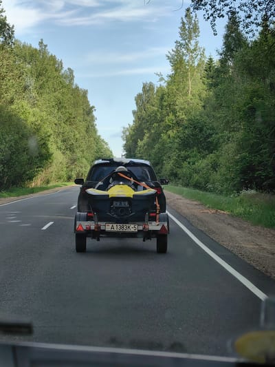 A black car with a trailer carrying a yellow jet ski drives on a paved road lined with trees. The license plate on the trailer reads "A 1383K-5". The sky is blue with some white clouds. The scene appears to be on a rural road, possibly in the vicinity of Хаценчыцкі сельскі Савет, Belarus.