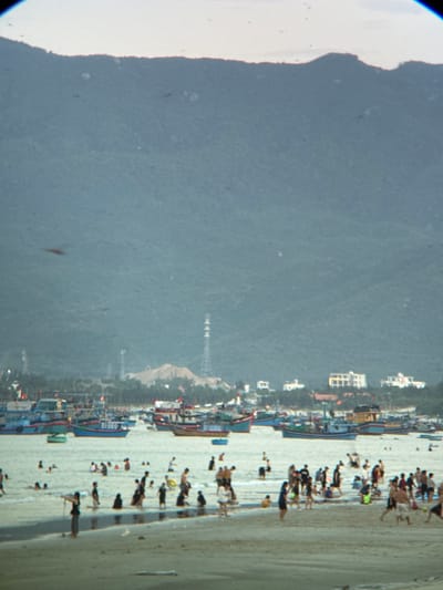 This image depicts a bustling beach scene in Dong Ninh Hoa, Vietnam. In the foreground, numerous people are engaged in various activities on the sandy shore and in the shallow water. Several individuals are walking, standing, and interacting with each other. Children and adults are scattered across the beach, with some wading in the water and others standing on the sand.
The midground of the image is dominated by a body of water filled with a multitude of boats. These boats, of varying sizes and designs, appear to be fishing vessels, with some docked and others anchored. The water is calm and reflects the light, creating a shimmering effect.
In the background, the landscape rises sharply, forming a mountainous backdrop. The slopes of the mountains are covered in lush vegetation, and hints of buildings and structures can be seen nestled amongst the trees. Tall, slender communication towers, possibly for broadcasting or telecommunications, are also visible on the terrain. The overall impression is of a vibrant coastal community, with a blend of human activity and natural scenery. The sky above is overcast, with a pale, diffused light suggesting either early morning or late afternoon.