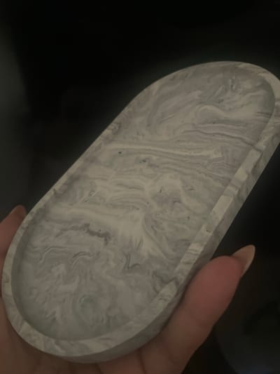 A hand holds an oval tray with a marble-like grey and white pattern. The tray is made of a solid material, possibly ceramic or concrete, and has a smooth, slightly glossy finish. The background is dark and out of focus, suggesting a dimly lit indoor environment. There are no discernible landmarks or contextual clues that would indicate a specific location within Torquay, United Kingdom. The image focuses solely on the tray and the hand holding it.