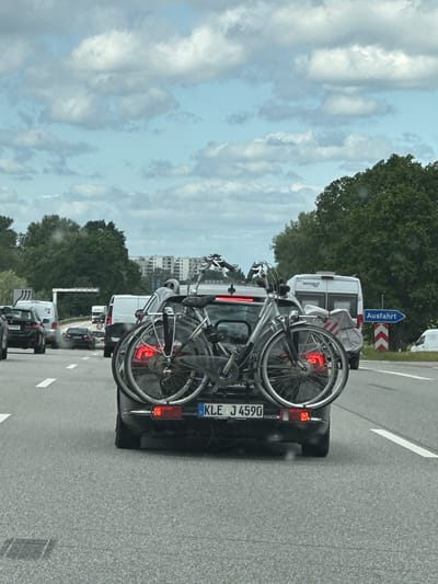 The image captures a traffic scene, likely on a German autobahn or highway. The focus is on a dark-colored car with a bicycle rack mounted on its rear, holding two bicycles. The car's license plate reads "KLE J 4590". The car is positioned in a line of other vehicles, indicating a potentially congested traffic situation. In the background, an "Ausfahrt" (exit) sign suggests proximity to a highway off-ramp. A white van is also visible ahead. The environment is characterized by trees lining the roadside, and the sky is overcast with cloudy conditions. Given the presence of a German license plate and the "Ausfahrt" sign, the location is likely within Germany. Without further information, a specific location in the city of Seevetal, Germany, cannot be pinpointed.